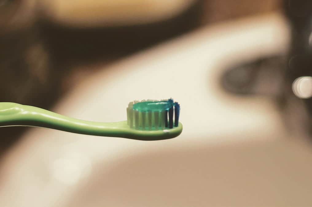 Grønn tannbørste med blått og grønt tannkrem på børstehodet, nærbilde foran uklart baderom. Daglig tannhygiene og forebyggende tannpleie.