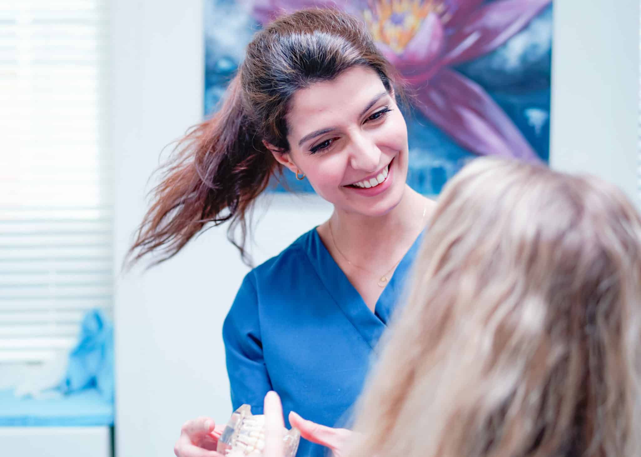 Kvinnelig tannlege i blå uniform smiler og viser pasient tannmodell. Konsultasjon om tannimplantat eller tannregulering i moderne klinikk.