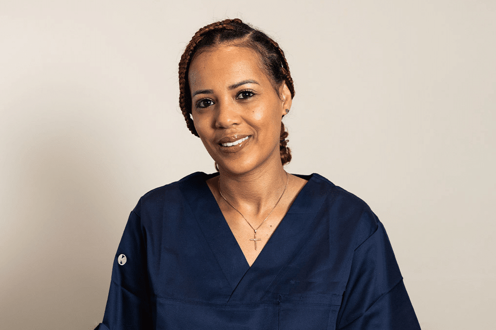 Kvinnelig helsearbeider med flettet hår og marineblå uniform smiler foran beige bakgrunn. Portrett av tannlege, tannpleier eller sykepleier i klinisk miljø.