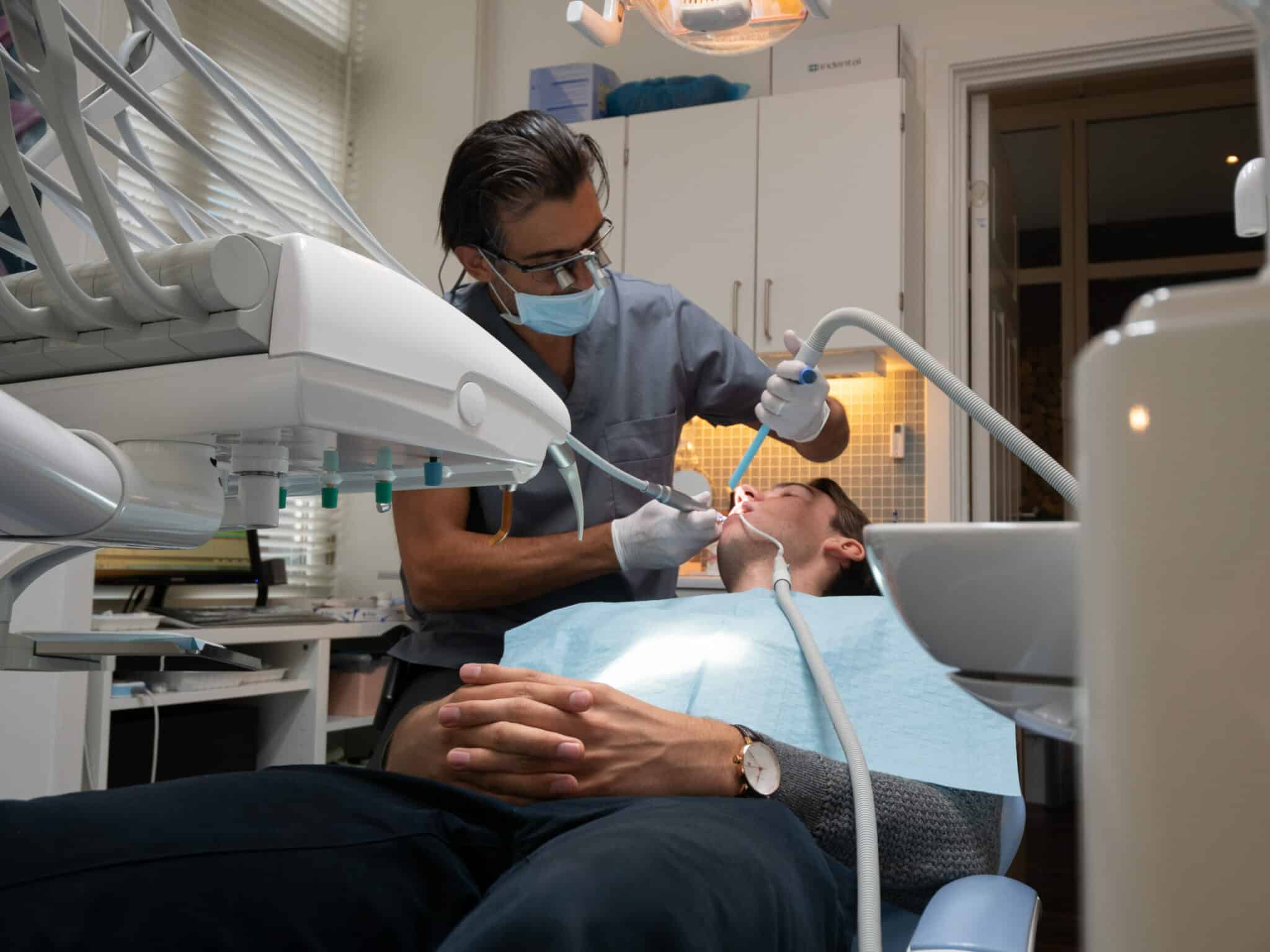 Tannlege utfører tannbehandling på mannlig pasient i moderne tannlegestol, med fokus på teknisk utstyr og profesjonell behandling.