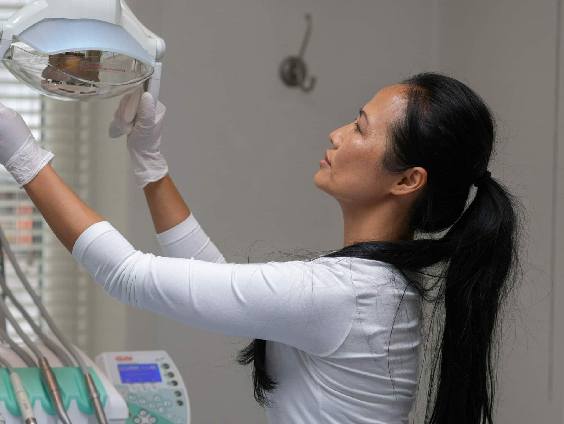 En kvinnelig tannpleier eller assistent i hvit overdel og hansker justerer den sterke operasjonslampen over behandlingsstolen for å sikre optimalt arbeidslys.