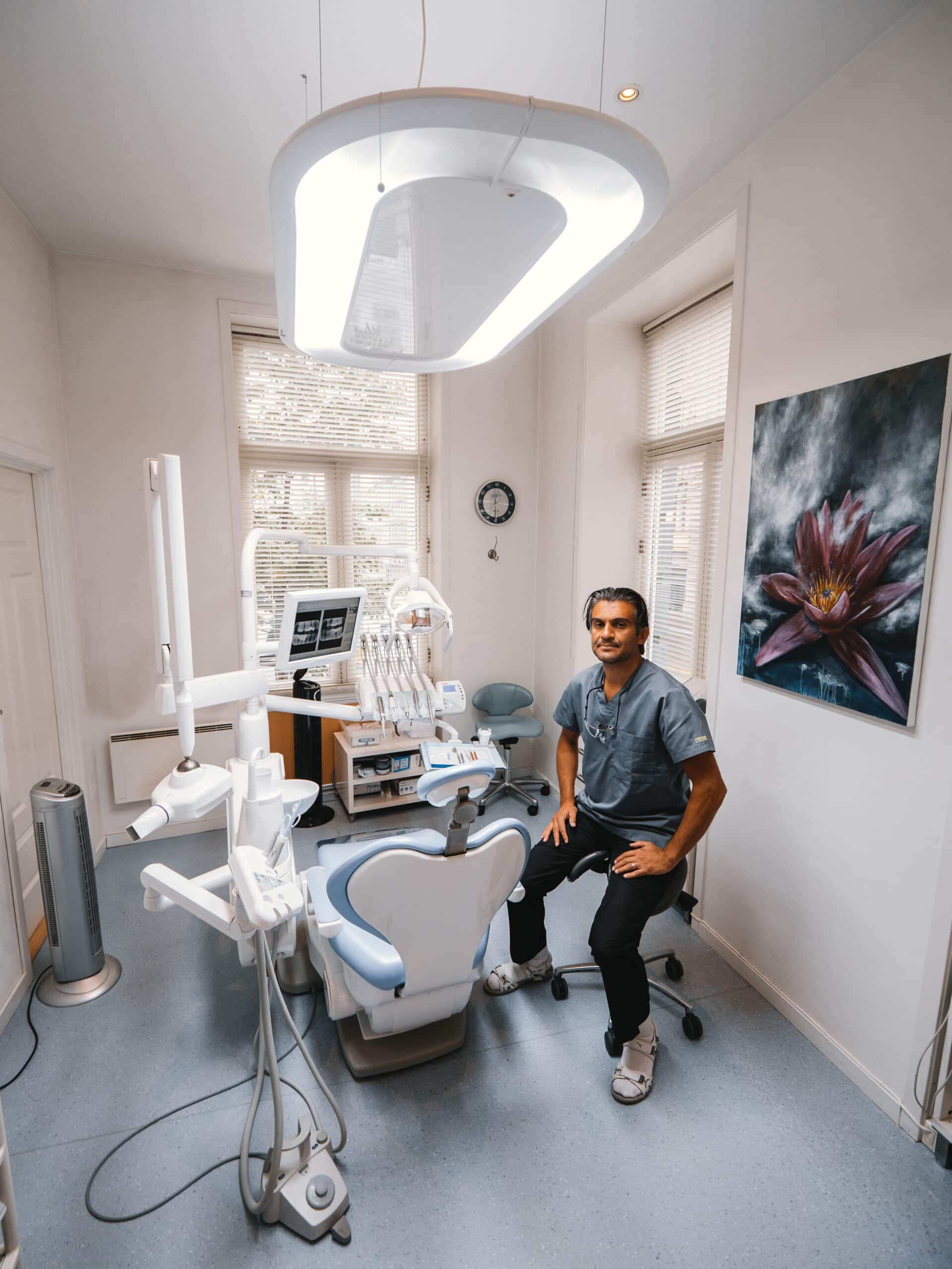 En mannlig tannlege i grå bekledning sitter ved siden av en moderne pasientstol i et lyst kontor med klassiske vinduer og kunst på veggene.
