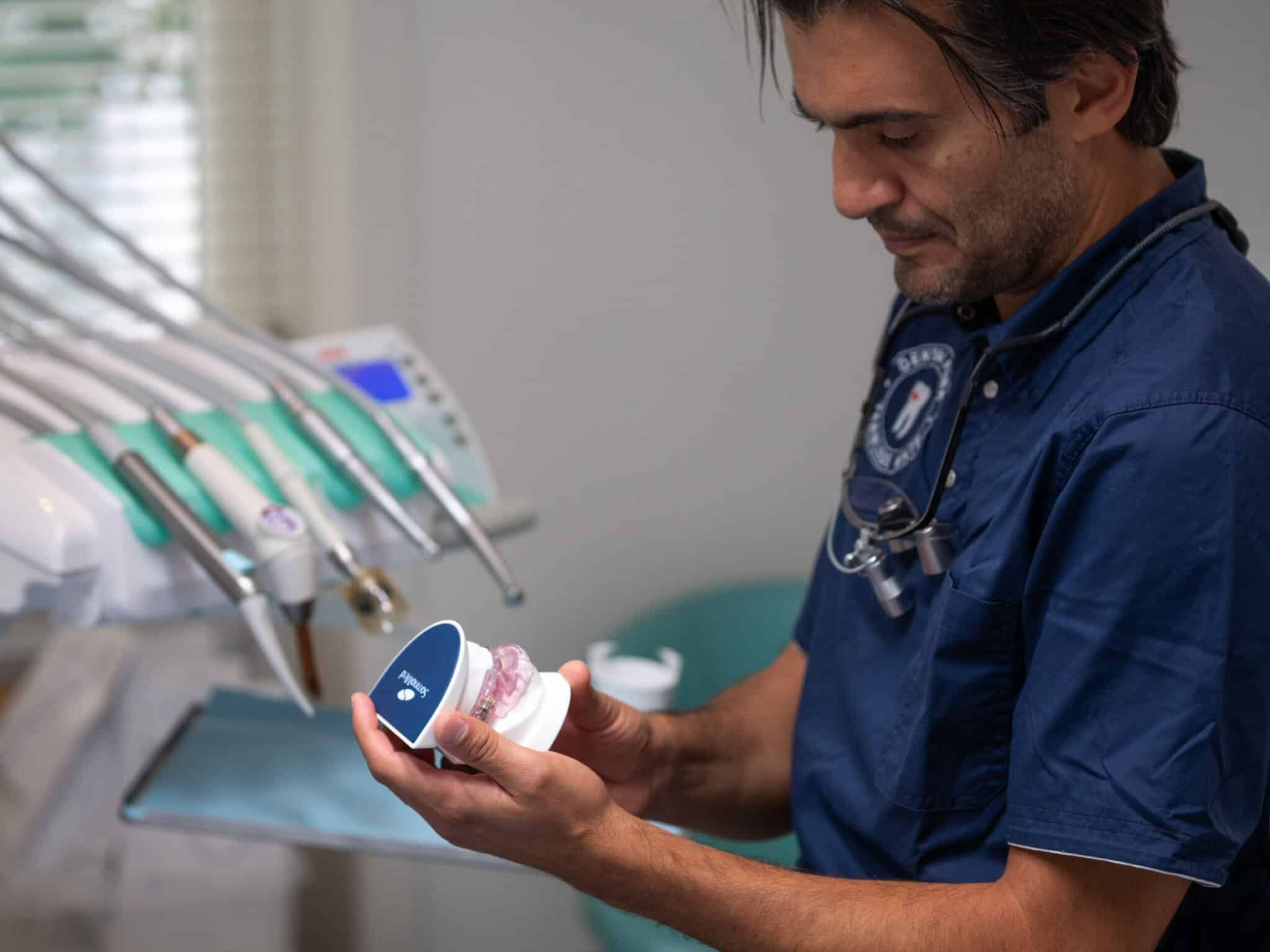 Nærbilde av en tannlege som holder en modell av en tanngard med en rosa bittskinne eller kjeveortopedisk apparat fra SomnoMed. Han studerer modellen nøye i et klinisk miljø.