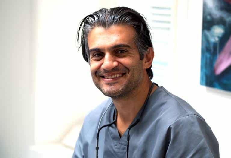 Mannlig tannlege eller lege med grå arbeidsklær og stetoskop rundt halsen smiler i lys klinikk. Portrett av helsearbeider i moderne tannlegekontor.