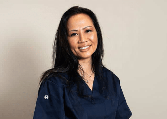 Et smilende portrett av en kvinne med langt mørkt hår, iført en profesjonell mørkeblå klinikkoverdel mot en nøytral, lys bakgrunn.