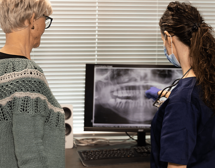 En kvinnelig tannlege i blå bekledning forklarer et røntgenbilde på en skjerm til en voksen kvinnelig pasient med briller og grønn genser. Begge står foran dataskjermen på et tannlegekontor.