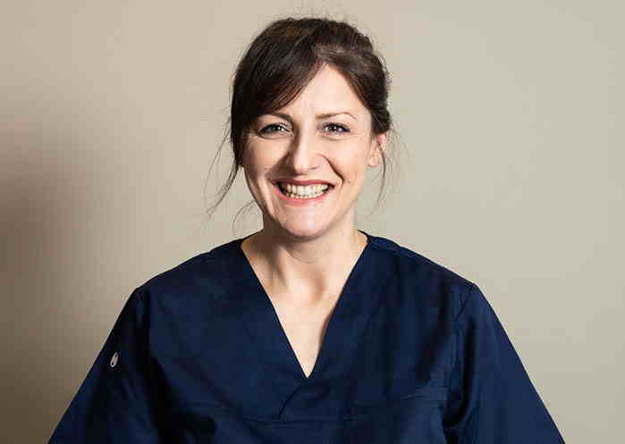 Kvinnelig helsearbeider i marineblå uniform smiler mot kamera foran en beige bakgrunn. Portrett av tannlege eller sykepleier i klinisk bekledning.