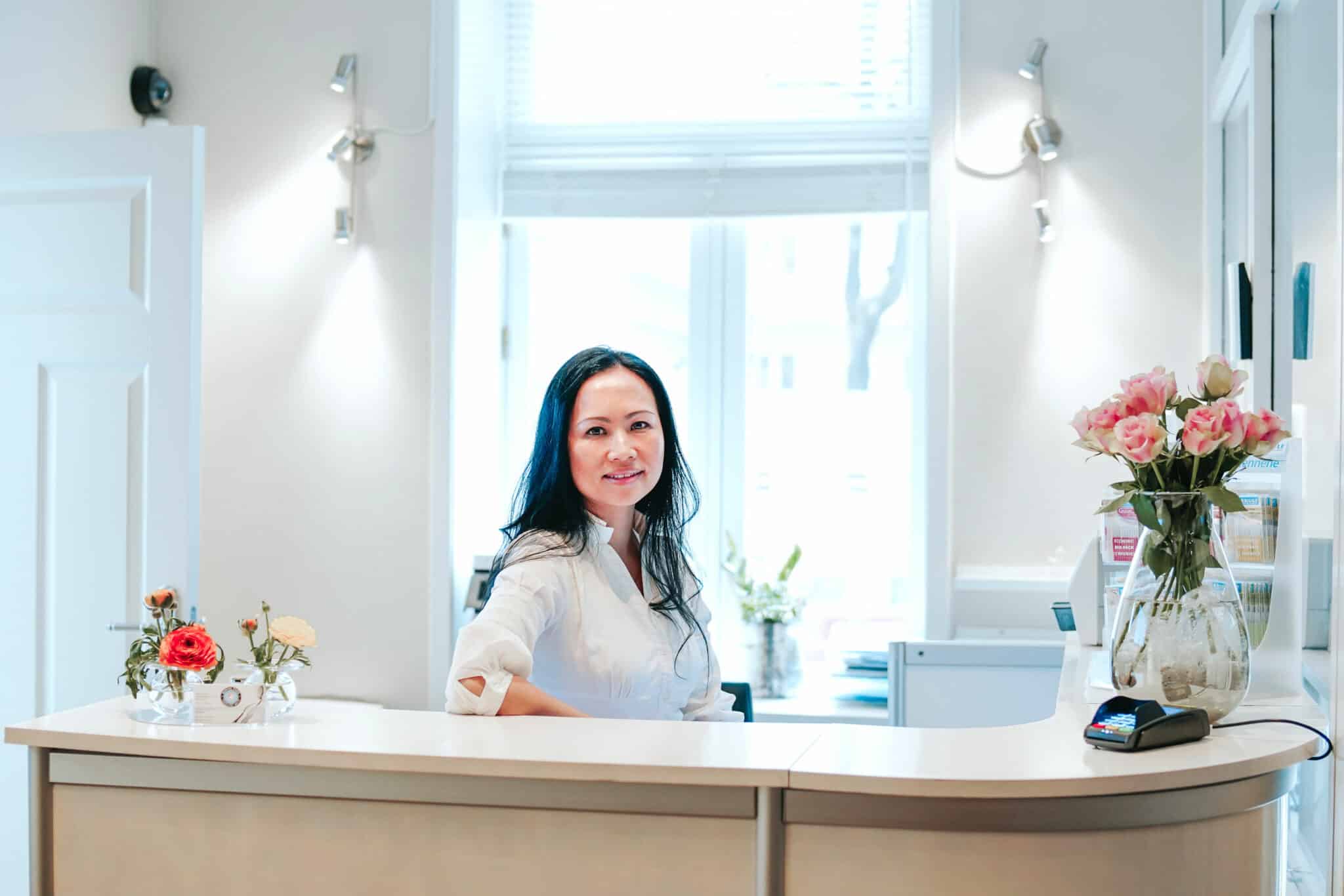 En kvinne med mørkt hår smiler fra bak en lys resepsjonsdisk dekorert med friske blomster i en lys klinikk.